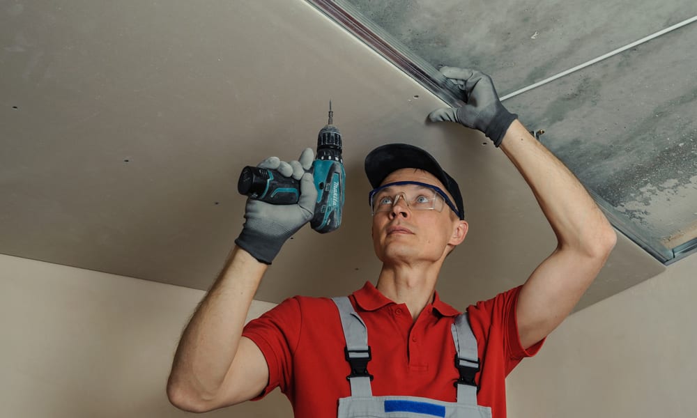 A workman holding up a Drywall board while screwing it in place. 